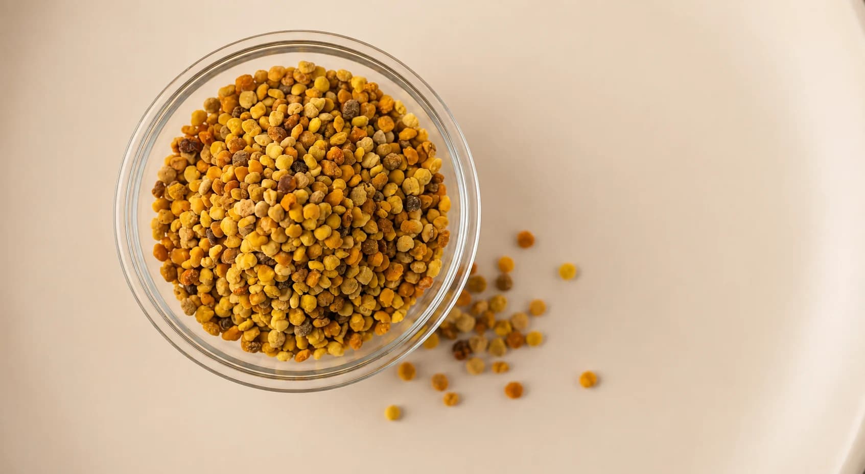 Loose bee pollen granules in a glass dish on a warm cream surface