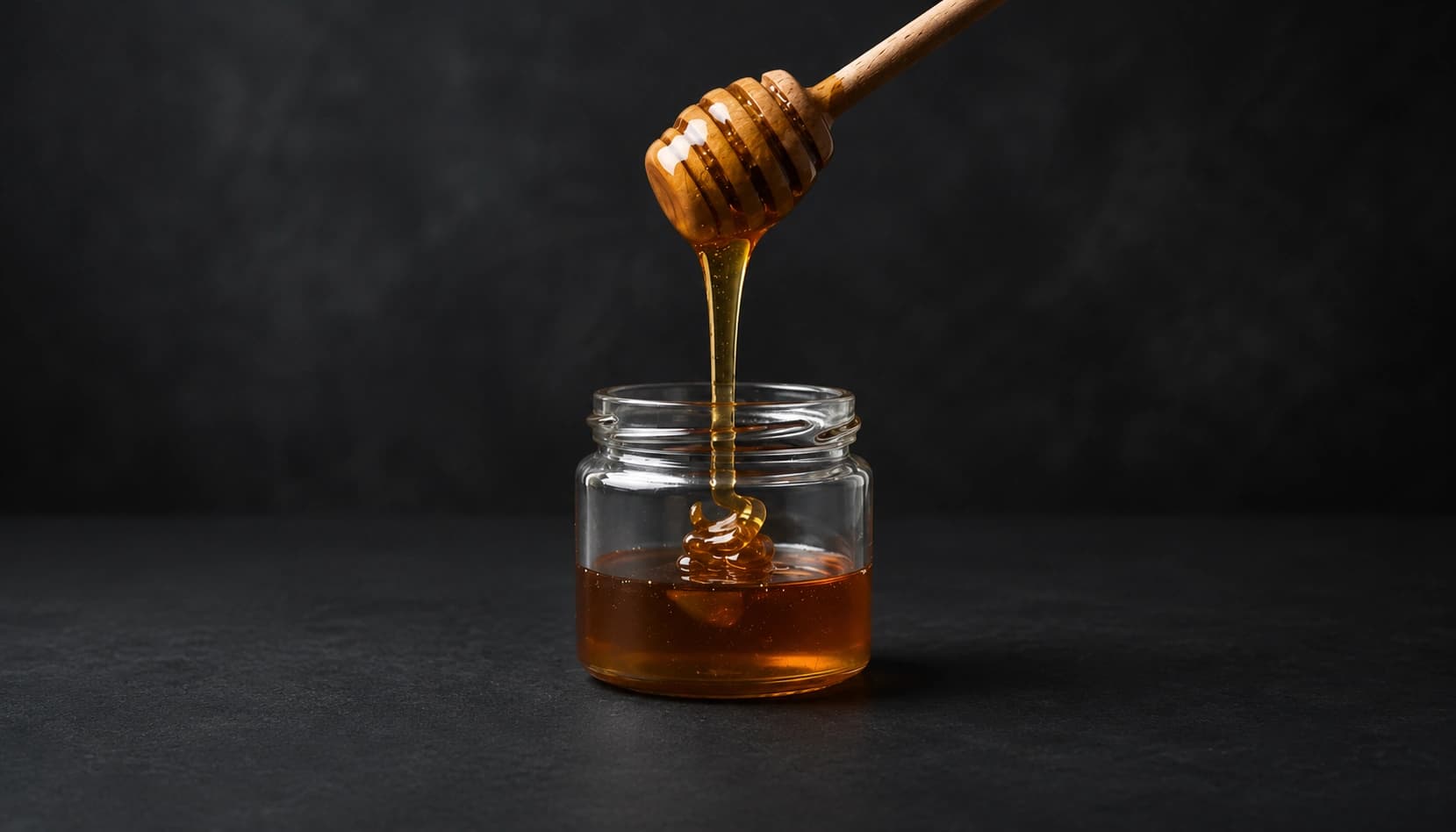 Raw honey being poured from a wooden dipper into a glass jar, catching the light against a dark background